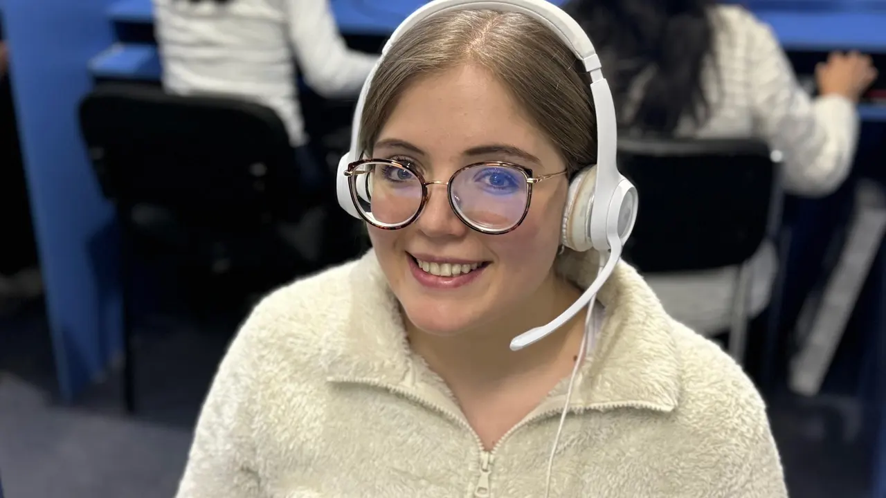 A human agent wearing a white headset smiles toward the camera in a call center, with colleagues at their desks behind her — call routing software