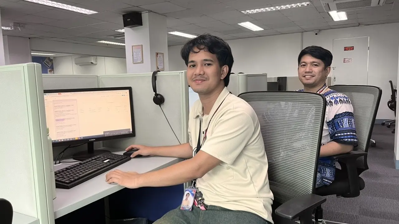 Two support agents sit at neighbouring partitioned desks on a contact center floor, one with his hands on the keyboard — customer service outsourcing