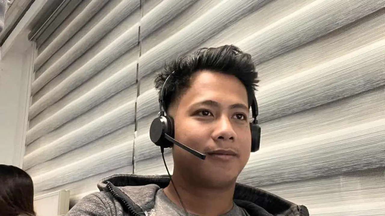 A support agent wearing a headset with a boom microphone, seen close up in front of window blinds — live chat outsourcing