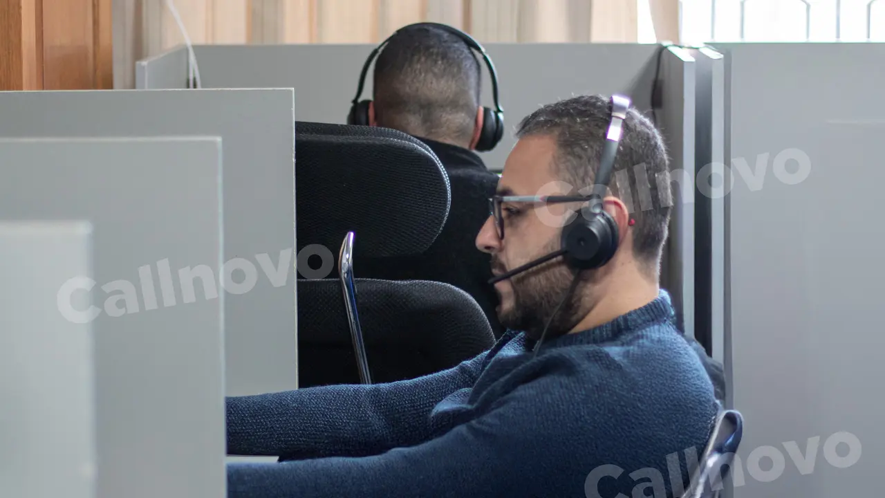 A support agent wearing a headset takes a call at a partitioned desk, seen from the side, with a colleague working behind him — outsourced IT support services