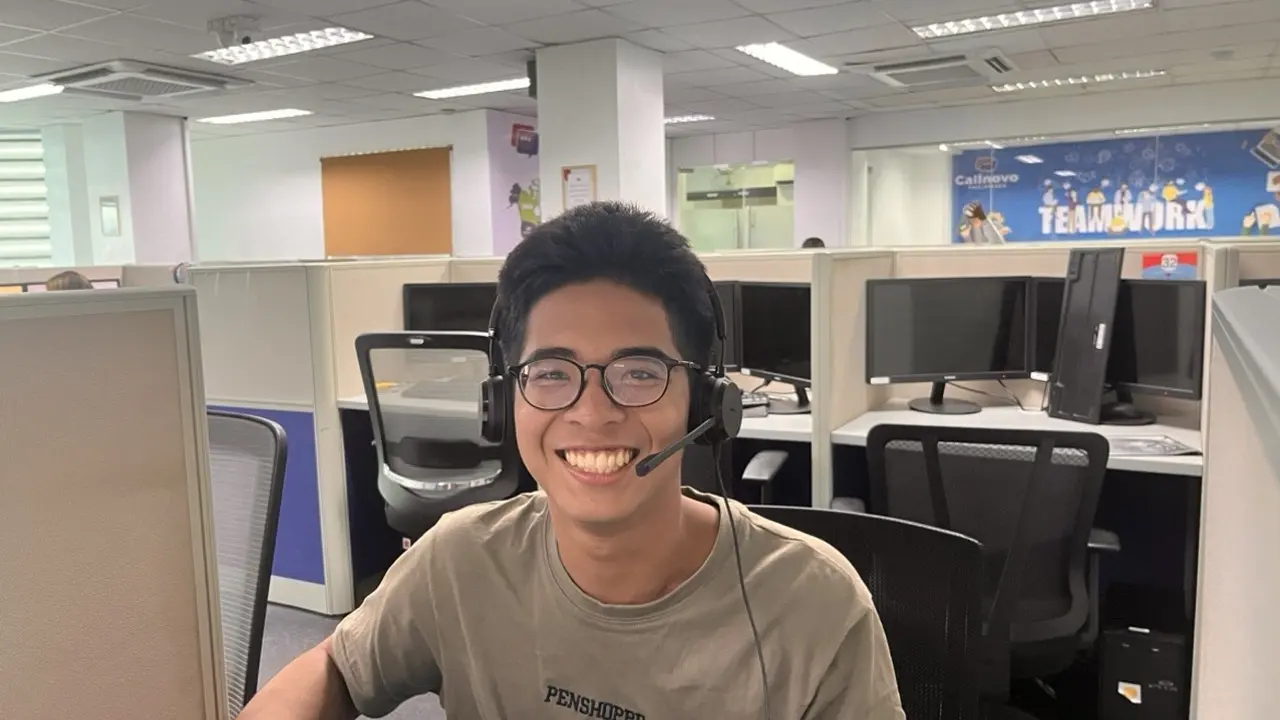 Un agente con auriculares sonríe a la cámara en una sala de call center, con puestos y monitores detrás — software de call center