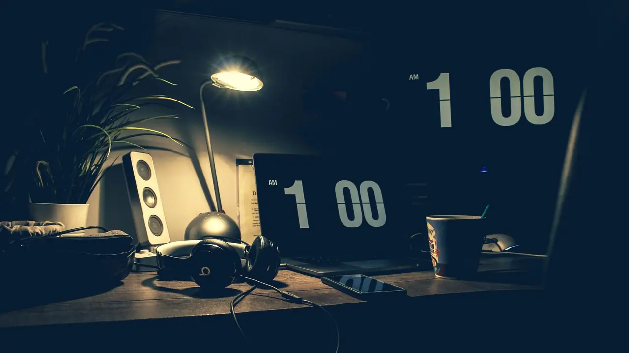 A dark home-office desk at 1:00 AM with a laptop, lamp, headphones and coffee — customer emails arriving while the support team is asleep