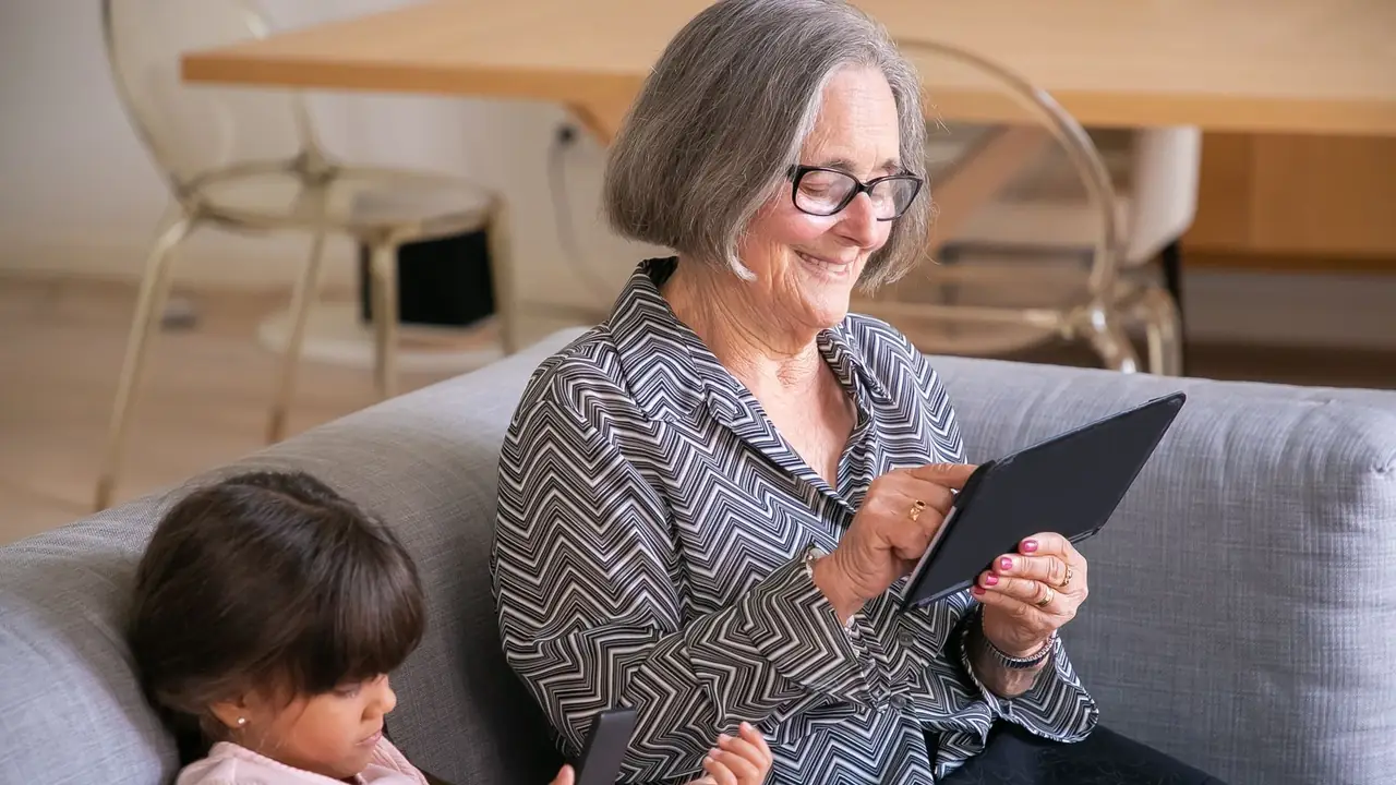 A senior woman smiling as she uses a tablet at home, her granddaughter beside her on the sofa