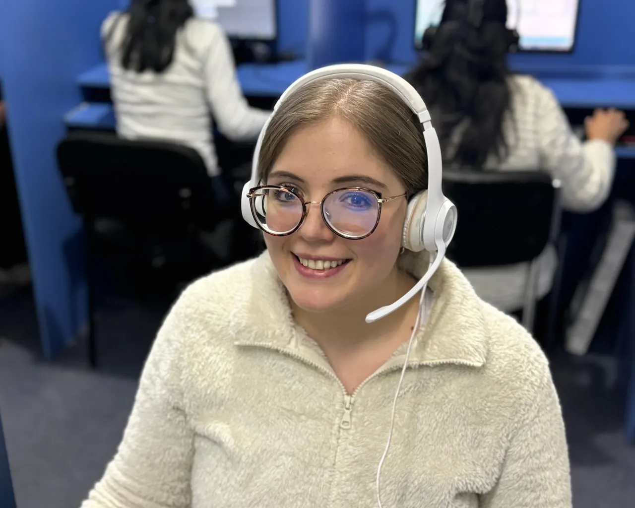 A Callnovo support agent wearing a headset smiles at her desk, with colleagues working behind her — the live team behind the AI