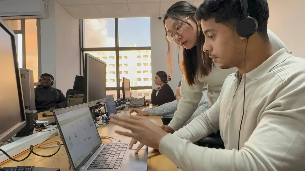 A colleague assists a headset-wearing agent at a computer workstation.