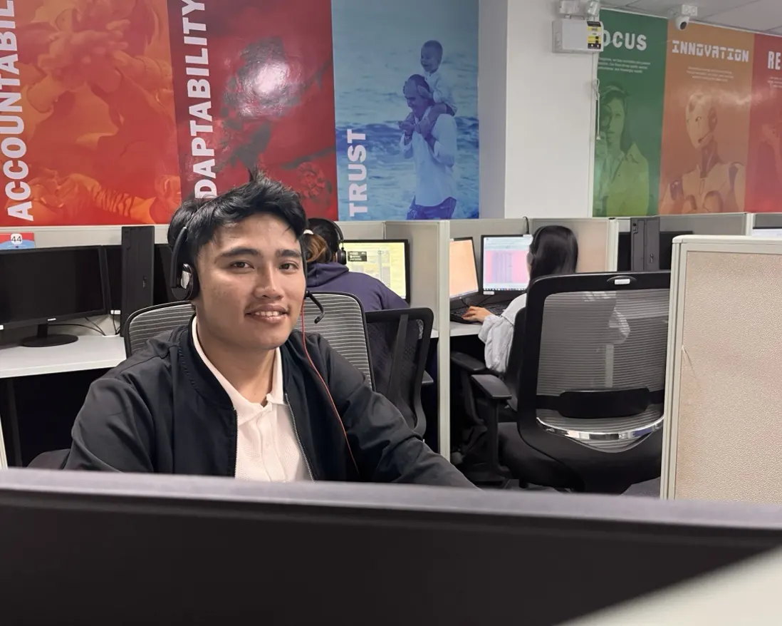 A Callnovo support agent wearing a headset smiles at his desk, with colleagues working behind him